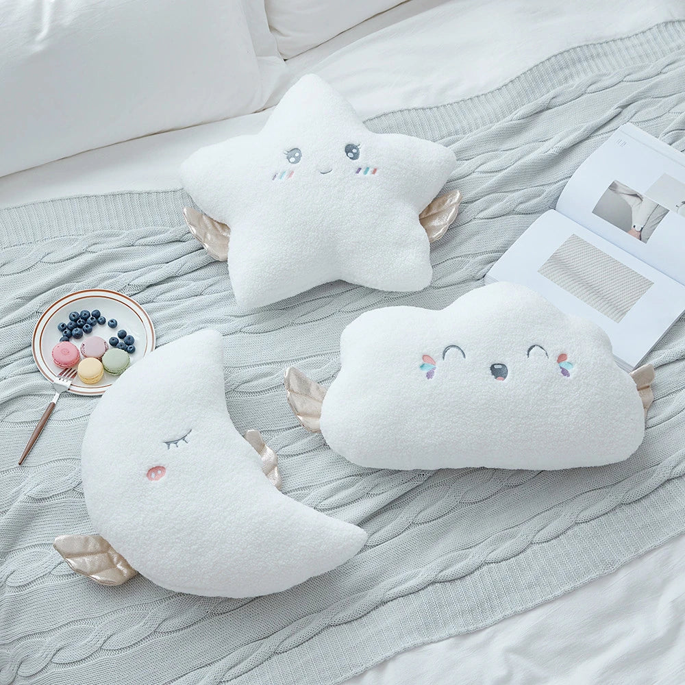 white moon, cloud, and star cushions with golden wings and pastel eyes and mouths on a bed