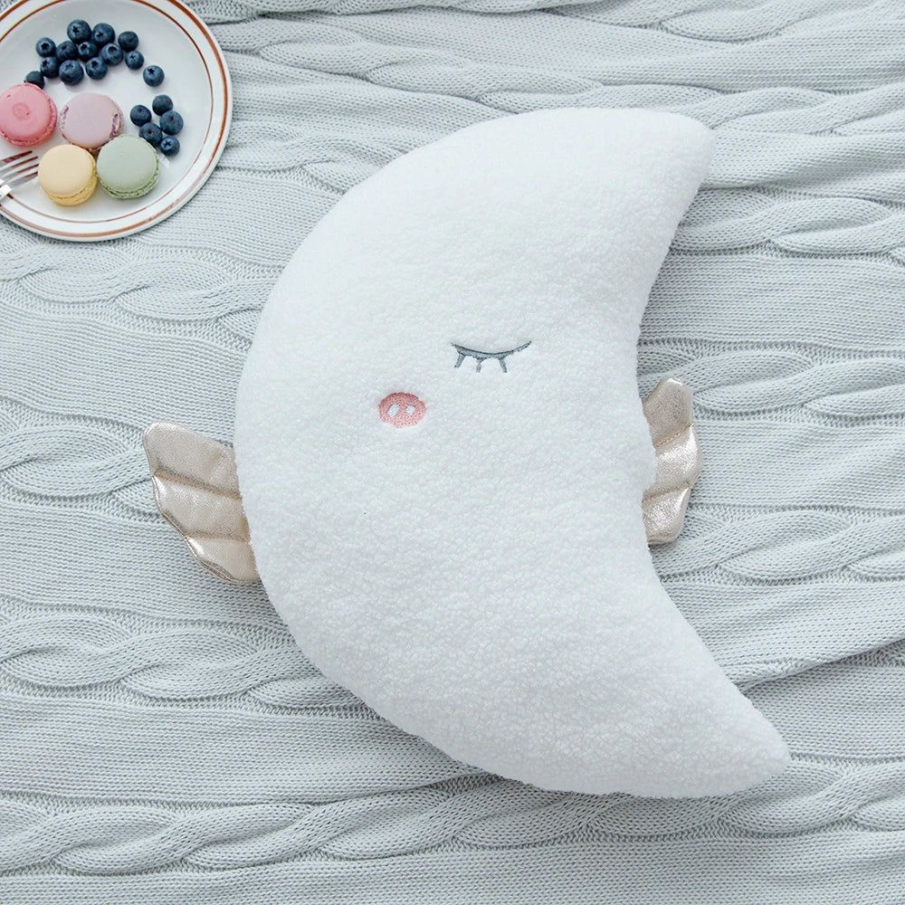 a white moon cushion with golden wings and pastel eyes and mouths on a bed